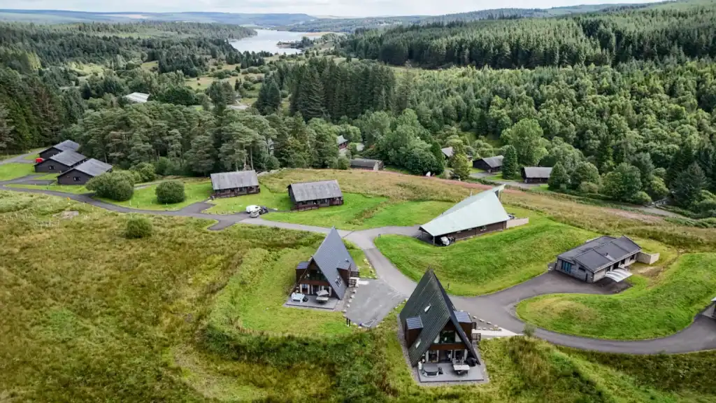 Aerial view of the Calvert Kielder site, showing timber A-frame lodges set among grassland and forest with Kielder Water in the distance